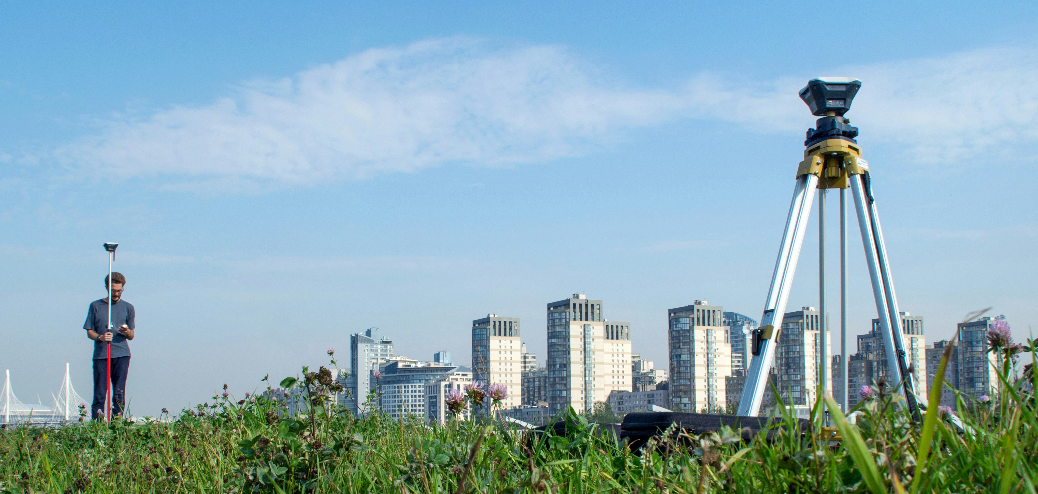 Ein Mann mit Vermessungsgerät auf einer Wiese. Im Hintergrund sieht man die Skyline einer Stadt.